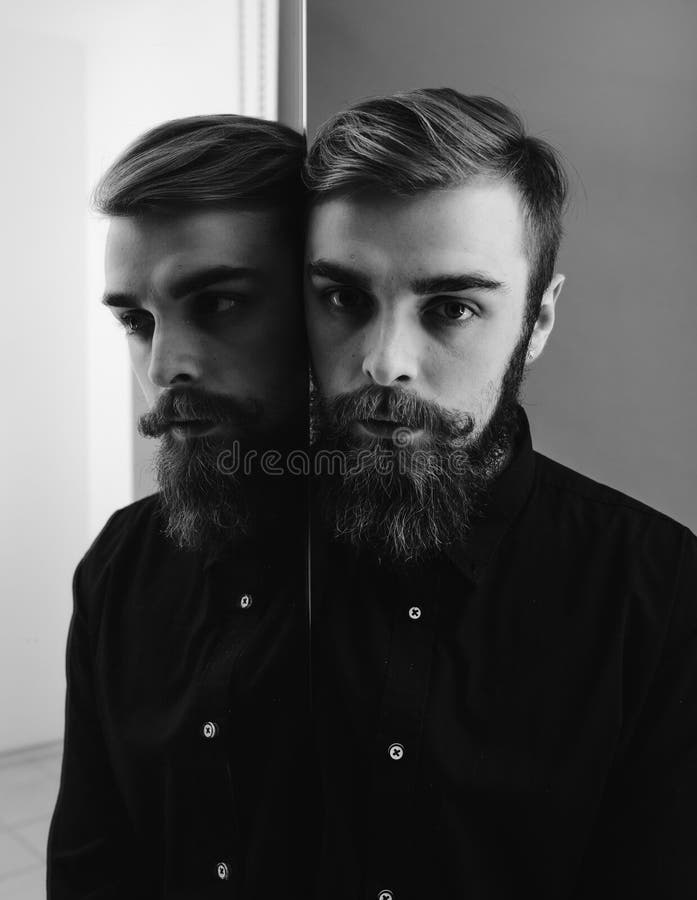 Svartvitt foto av en man med ett sk?gg och en stilfull frisyr som ?r ikl?dda det svarta skjortaanseendet bredvid spegeln royaltyfria bilder Svartvitt foto av en man med ett sk?gg och en stilfull frisyr som ?r ikl?dda det svarta skjortaanseendet bredvid spegeln royaltyfria bilder