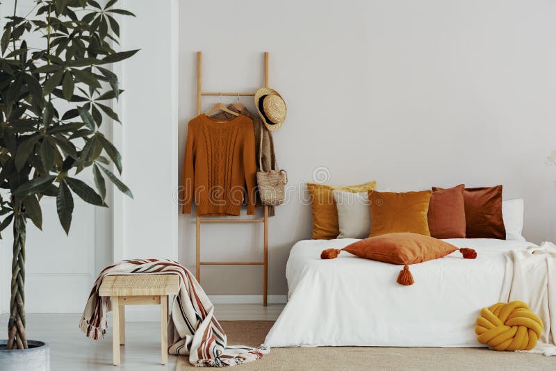 Sweaters, wicker hat and bag on wooden ladder next to king size bed with velvet pillows, copy space on empty wall