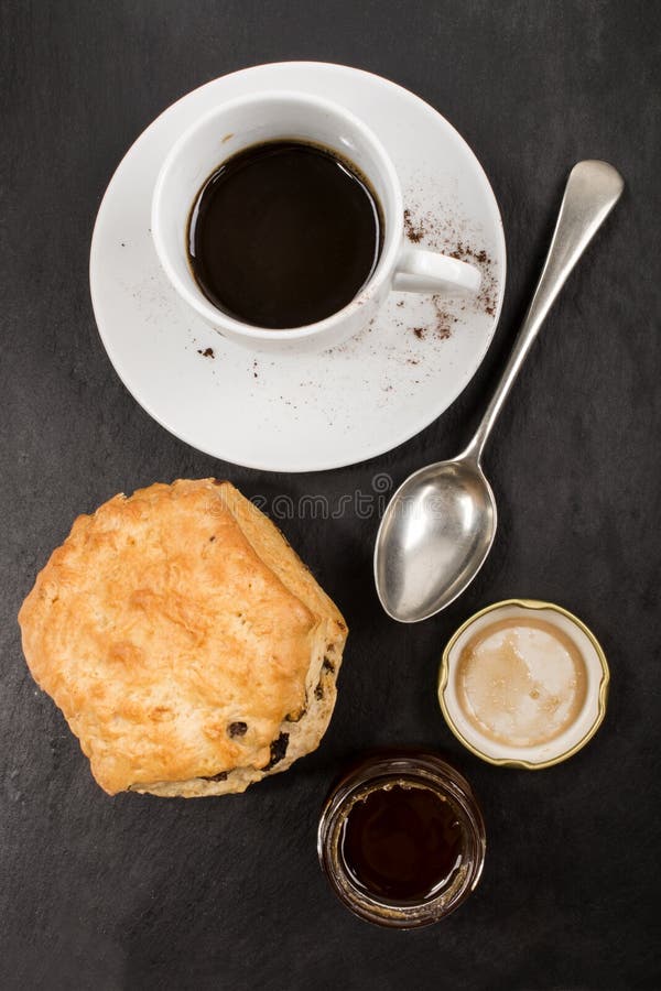 Sweet british raisin scone and a glass filled with honey, cup hot instant coffee also small spoon on slate