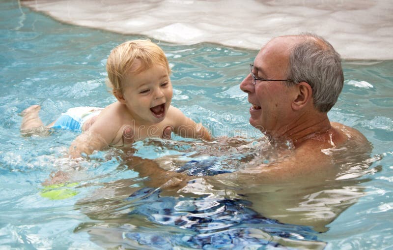 Muž s strieborné vlasy a okuliare drží šťastný jeden rok staré dieťa, chlapca s blond vlasy a modré oči v bazéne.