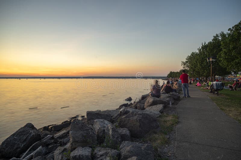 SYLVAN BEACH, NEW YORK - JULY 3, 2019: Sunset at Sylvan Beach of the Oneida lake while people waiting for the Independence Day Fireworks, sand, upstate, sea, walking, kids, holiday, family, happiness, shoreline, travel, madison, county, coast, swimming, summer, play, water, woman, joy, parents, entertainment, lifestyle, fun, verona, village, together, outdoor, vacation, active, waves, ny, weekend, outside, tide, healthy, leisure, happy, kiosk, crowd, having, fluffy, young, love, noon, exotic. SYLVAN BEACH, NEW YORK - JULY 3, 2019: Sunset at Sylvan Beach of the Oneida lake while people waiting for the Independence Day Fireworks, sand, upstate, sea, walking, kids, holiday, family, happiness, shoreline, travel, madison, county, coast, swimming, summer, play, water, woman, joy, parents, entertainment, lifestyle, fun, verona, village, together, outdoor, vacation, active, waves, ny, weekend, outside, tide, healthy, leisure, happy, kiosk, crowd, having, fluffy, young, love, noon, exotic