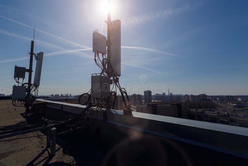 Silhouette of of Panel antenna of GSM DCS UMTS LTE bands and radio unit are as part of communication equipment of basic station are installed on the roof and sky with sun and city are as background. Silhouette of of Panel antenna of GSM DCS UMTS LTE bands and radio unit are as part of communication equipment of basic station are installed on the roof and sky with sun and city are as background.