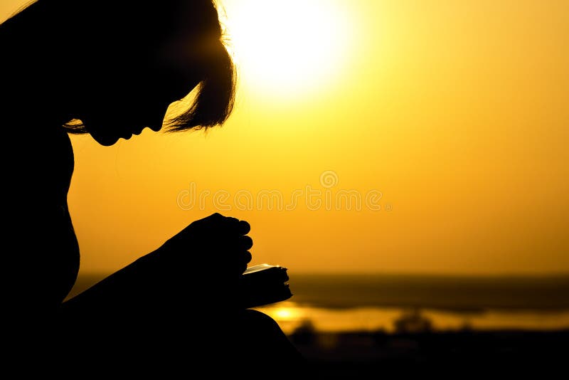 Silhouette of young woman bowed her head and praying to God in the nature, the girl holds her hands on the Bible near the tree at sunset, the concept of religion and spirituality. Silhouette of young woman bowed her head and praying to God in the nature, the girl holds her hands on the Bible near the tree at sunset, the concept of religion and spirituality