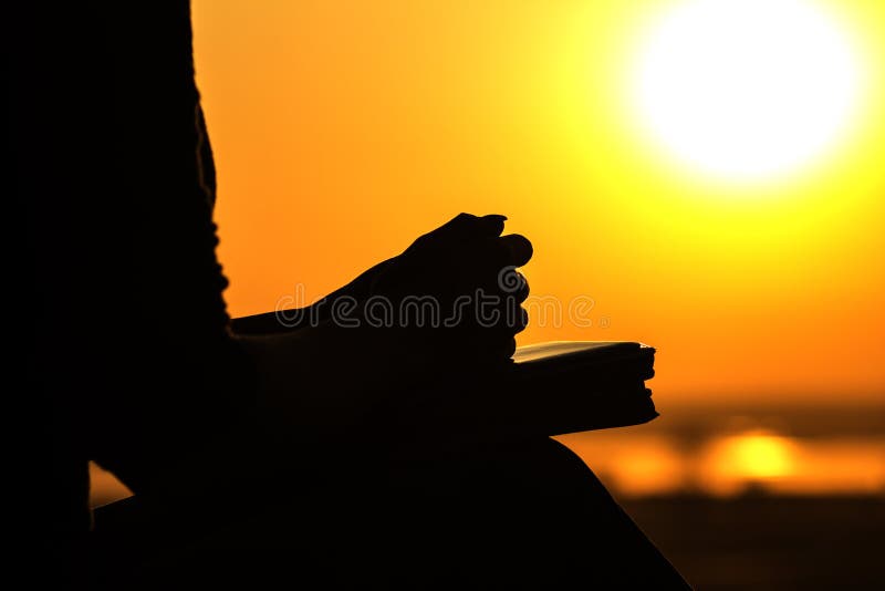 Silhouette of the hands of a young woman praying to God in the nature, the girl holds her hands on the Bible near the tree at sunset, the concept of religion and spirituality. Silhouette of the hands of a young woman praying to God in the nature, the girl holds her hands on the Bible near the tree at sunset, the concept of religion and spirituality
