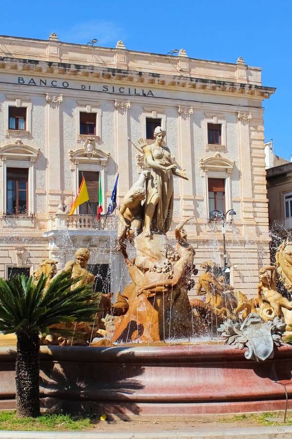 Syracuse, Sicily, Italy - Apr 10th 2019: Amazing Fountain of Diana on the Archimedes Square in beautiful Ortigia Island. Building of a Sicilian bank in the background. Sunny day, blue sky. Syracuse, Sicily, Italy - Apr 10th 2019: Amazing Fountain of Diana on the Archimedes Square in beautiful Ortigia Island. Building of a Sicilian bank in the background. Sunny day, blue sky