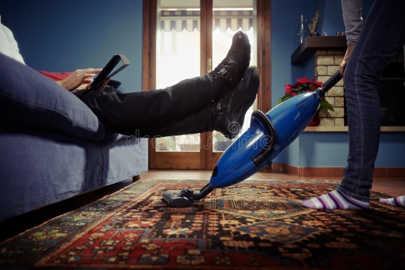 Caucasian heterosexual couple, with woman doing chores using vacuum cleaner on carpet and lazy man on sofa with tablet pc. Horizontal shape, low angle view. Caucasian heterosexual couple, with woman doing chores using vacuum cleaner on carpet and lazy man on sofa with tablet pc. Horizontal shape, low angle view