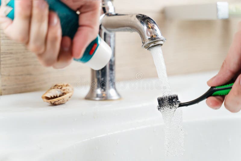 A toothbrush and toothpaste in the hand of a man under a stream of water that flows from under the faucet. A toothbrush and toothpaste in the hand of a man under a stream of water that flows from under the faucet