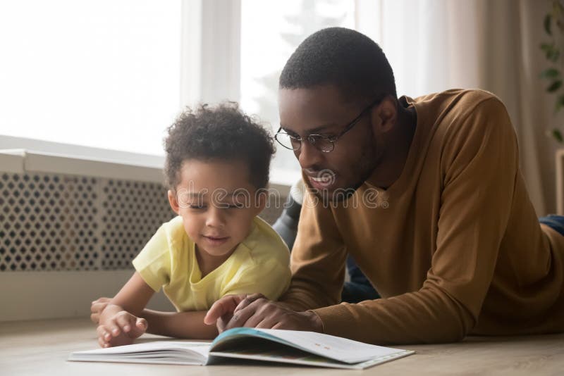 Happy black father baby sitter and toddler son having fun with book lying on warm floor, african dad teaching little kid boy learning to read, parent with children educational activities at home. Happy black father baby sitter and toddler son having fun with book lying on warm floor, african dad teaching little kid boy learning to read, parent with children educational activities at home