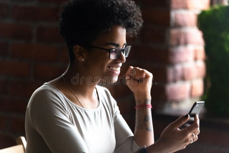 Happy smiling African American woman using phone, celebrate received good news with gesture, excited female looking at smartphone screen, browsing mobile apps, reading email, online game win. Happy smiling African American woman using phone, celebrate received good news with gesture, excited female looking at smartphone screen, browsing mobile apps, reading email, online game win