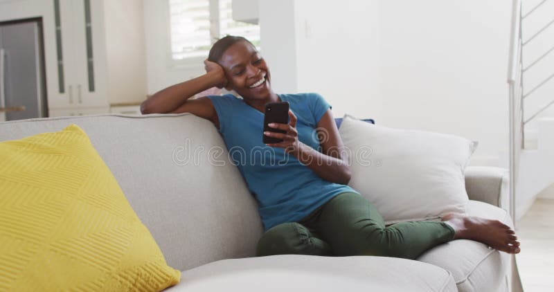 Szczęśliwa amerykańska matka afrykańska siedząca na sofie przy użyciu smartfona i śmiejąca się