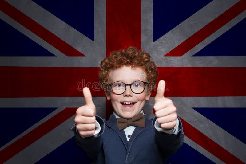 Szczęśliwa dziecko chłopiec pokazuje kciuk w górę UK chorągwianego tła przeciw Uczy się angielszczyzny swój chłodno zdjęcia stock Szczęśliwa dziecko chłopiec pokazuje kciuk w górę UK chorągwianego tła przeciw Uczy się angielszczyzny swój chłodno zdjęcia stock