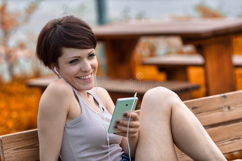 Happy young woman smiling with pleasure as she listens to music on earbuds using her smartphone while relaxing outdoors on a park bench. Happy young woman smiling with pleasure as she listens to music on earbuds using her smartphone while relaxing outdoors on a park bench