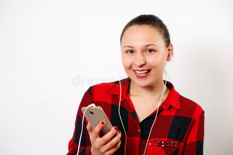 Szczęśliwa młoda piękna dziewczyna słucha muzyka w hełmofonach i ono uśmiecha się w czerwonej szkockiej kraty koszula Na bia?ym t obraz royalty free Szczęśliwa młoda piękna dziewczyna słucha muzyka w hełmofonach i ono uśmiecha się w czerwonej szkockiej kraty koszula Na bia?ym t obraz royalty free
