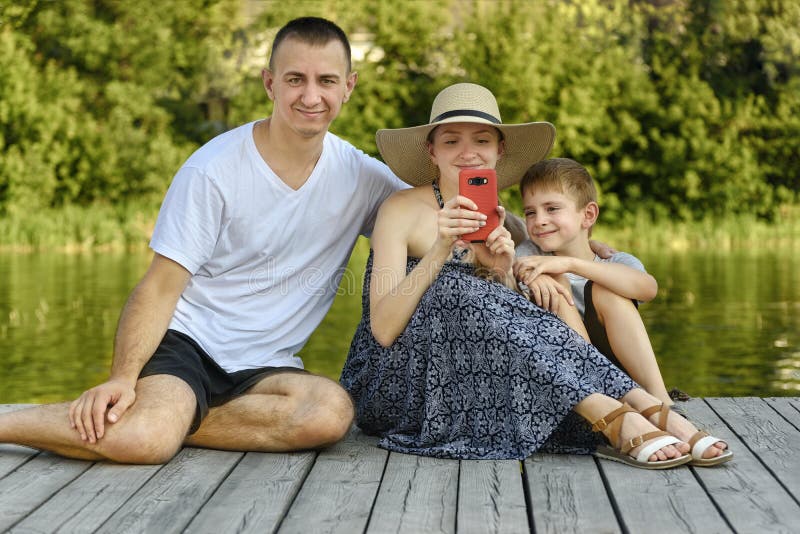 Szczęśliwa młoda rodzina, ojciec matka i dwa małego syna, jesteśmy siedzący selfies na rzecznym molu i brać obrazy stock Szczęśliwa młoda rodzina, ojciec matka i dwa małego syna, jesteśmy siedzący selfies na rzecznym molu i brać obrazy stock