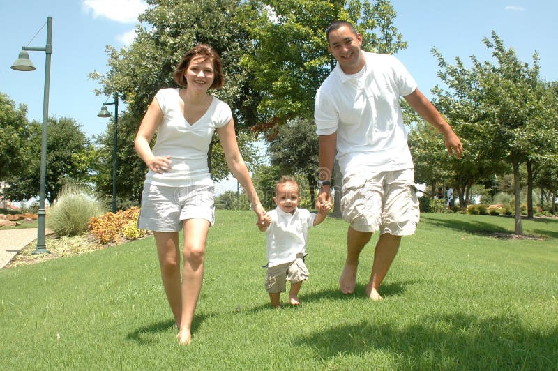 Young married couple with their toddler child. New family taking pictures in white shirts and tan shorts. Family of three holding hands as they walk the park. Young married couple with their toddler child. New family taking pictures in white shirts and tan shorts. Family of three holding hands as they walk the park.