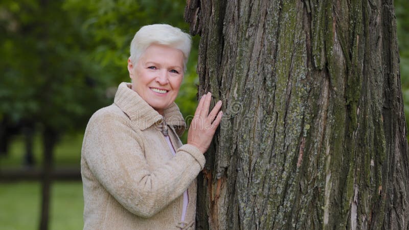 Szczęśliwa spokojna dojrzała samotna kobieta w średnim wieku patrząca na kamerę pozującą się dużego drzewa uśmiechającą się starsz