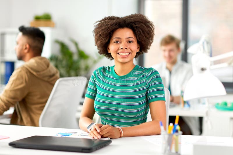 Business, education and people concept - happy smiling african american women with laptop computer sitting at office table. Business, education and people concept - happy smiling african american women with laptop computer sitting at office table