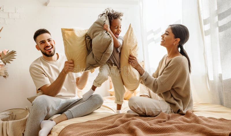 Szczęśliwa wieloetniczna matka i dziecko śmieją się bawiąc się w poduszki i skakając do łóżka w domu