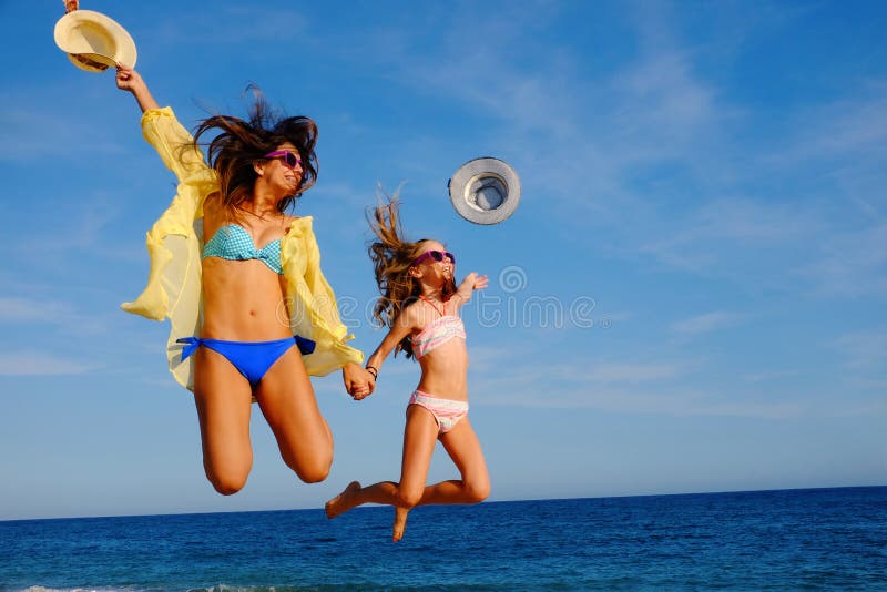 Action portrait of young mother with daughter jumping together on beach. Laughing girls in swimwear throwing hats in air. Action portrait of young mother with daughter jumping together on beach. Laughing girls in swimwear throwing hats in air.