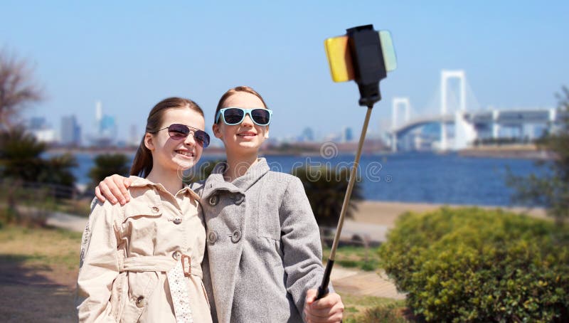 People, children, friends and technology concept - happy girls taking picture with smartphone on selfie stick over tokyo rainbow bridge in japan background. People, children, friends and technology concept - happy girls taking picture with smartphone on selfie stick over tokyo rainbow bridge in japan background