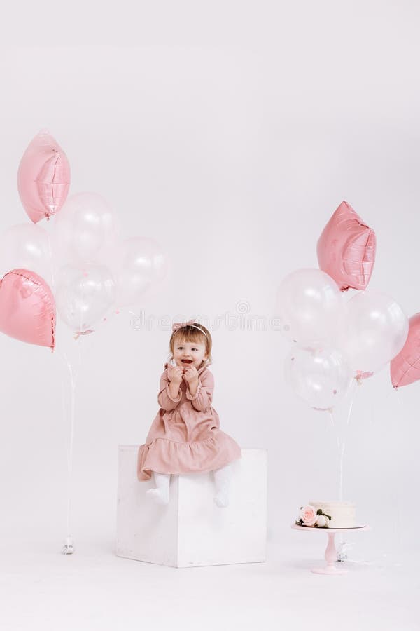 happy birthday 2 years old little girl in pink dress. white cake with candles and roses. Birthday decorations with white and pink color balloons and confetti for party on a white wall. Happy birthday. happy birthday 2 years old little girl in pink dress. white cake with candles and roses. Birthday decorations with white and pink color balloons and confetti for party on a white wall. Happy birthday.
