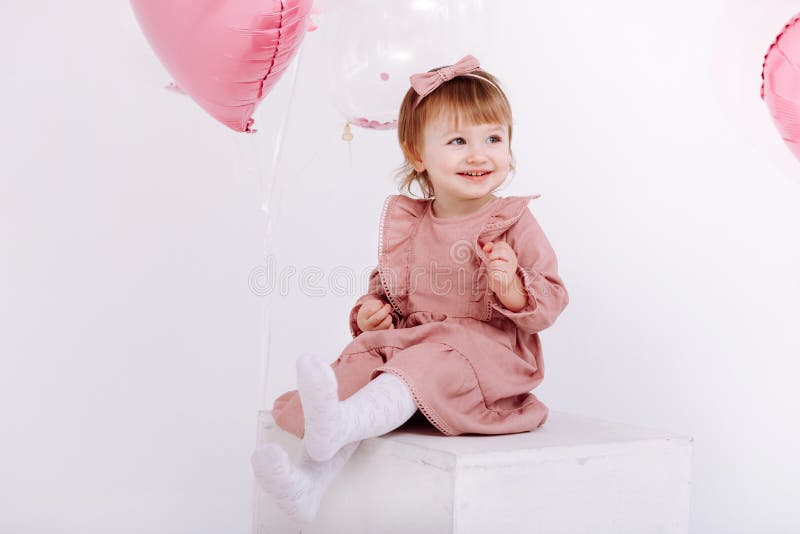 Happy birthday 2 years old little girl in pink dress. white cake with candles and roses. Birthday decorations with white and pink color balloons and confetti for party on a white wall. Happy birthday. Happy birthday 2 years old little girl in pink dress. white cake with candles and roses. Birthday decorations with white and pink color balloons and confetti for party on a white wall. Happy birthday.