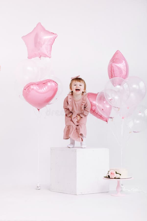 Happy birthday 2 years old little girl in pink dress. white cake with candles and roses. Birthday decorations with white and pink color balloons and confetti for party on a white wall. Happy birthday. Happy birthday 2 years old little girl in pink dress. white cake with candles and roses. Birthday decorations with white and pink color balloons and confetti for party on a white wall. Happy birthday.