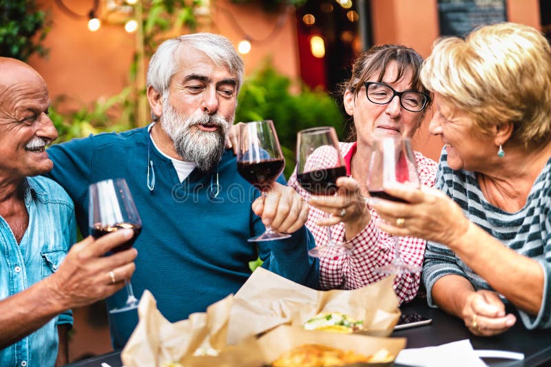 Szczęśliwi seniorzy, którzy bawią się tostując czerwone wino na kolacji w ogródku... emerytowane pary pijące razem w restauracji obraz stock