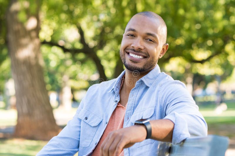 Young african man relaxing at park in a summer day. Happy black cheerful guy feeling good and sitting on bench at park. Smiling american man looking at camera outdoor. Young african man relaxing at park in a summer day. Happy black cheerful guy feeling good and sitting on bench at park. Smiling american man looking at camera outdoor.