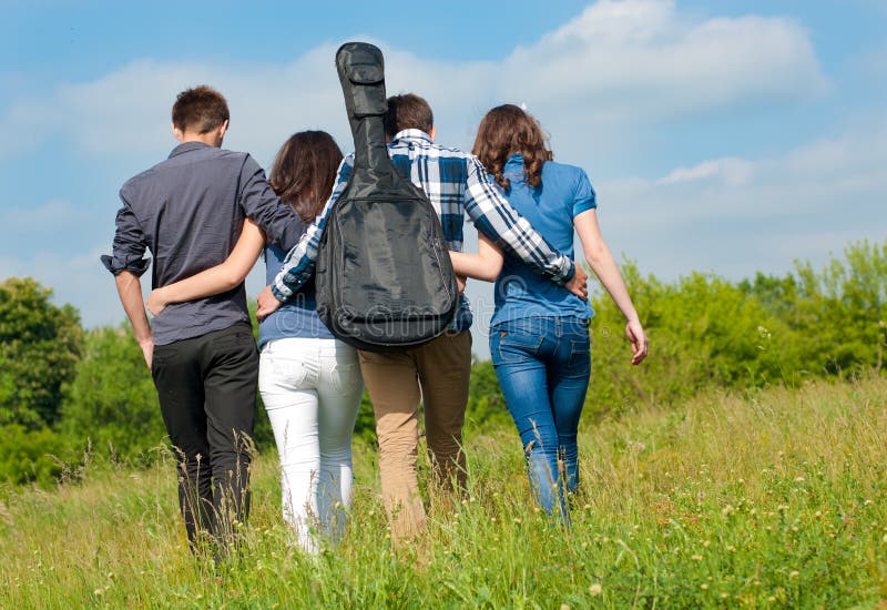 Happy mixed group of Young people two beautiful women and two handsome young men with guitar walking outdoors away from camera on a bright sunny day of spring or summer on the blue sky and green field background. Happy mixed group of Young people two beautiful women and two handsome young men with guitar walking outdoors away from camera on a bright sunny day of spring or summer on the blue sky and green field background