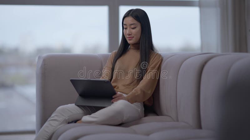 Szerokie zdjęcie czarującej azjatyckiej kobiety surfującej po Internecie na tablecie siedzącym na kanapie w biurze domowym. radosn