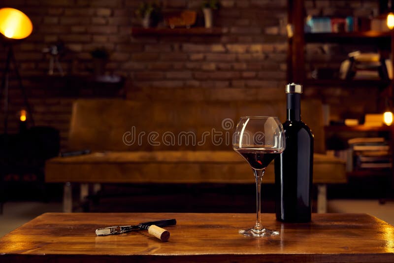 Wine glass and a bottle of red wine on table at home, cosy dark living room in background. Autumn mood with dark and warm colors. Wine glass and a bottle of red wine on table at home, cosy dark living room in background. Autumn mood with dark and warm colors.