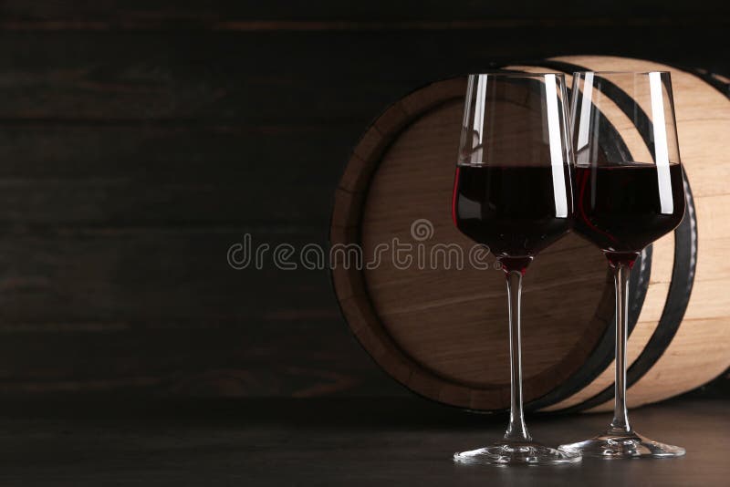 Glasses of red wine and wooden barrel on table against dark background. Space for text. Glasses of red wine and wooden barrel on table against dark background. Space for text