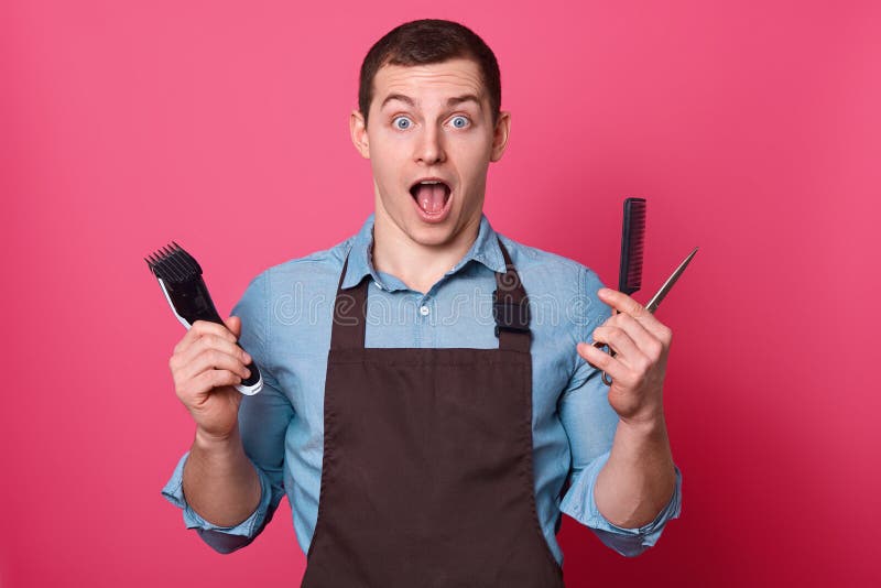 Shocked male barber holds necessary equipment for making hairstyle, keeps jaw dropped, stares at camera, wears shirt and apron, isolated over pink background. Omg, how many clients in barbershop. Shocked male barber holds necessary equipment for making hairstyle, keeps jaw dropped, stares at camera, wears shirt and apron, isolated over pink background. Omg, how many clients in barbershop