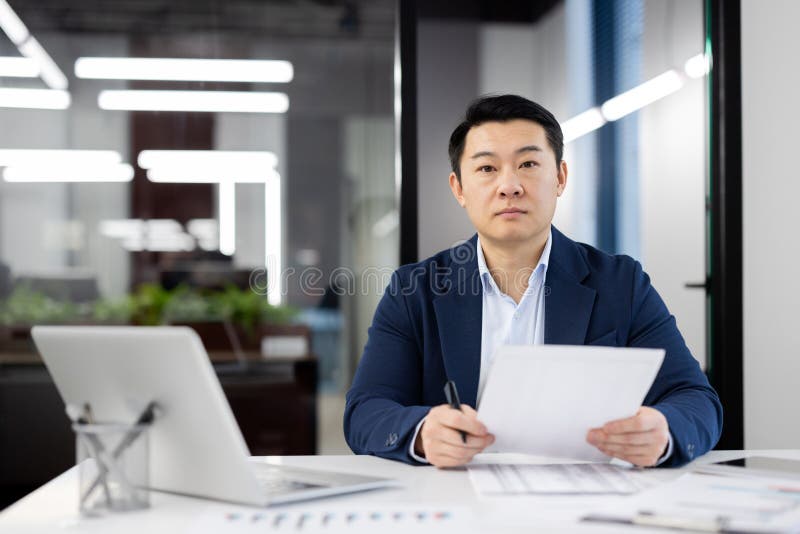 Focused Asian businessman sitting at desk in a well-lit office, reviewing documents with serious expression, displaying professionalism and determination. Focused Asian businessman sitting at desk in a well-lit office, reviewing documents with serious expression, displaying professionalism and determination.