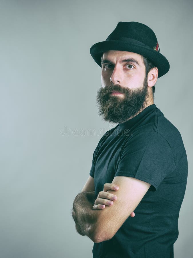 Säker hipster som bär den svarta hatten och t-skjortan som ser kameran med korsade armar fotografering för bildbyråer Säker hipster som bär den svarta hatten och t-skjortan som ser kameran med korsade armar fotografering för bildbyråer