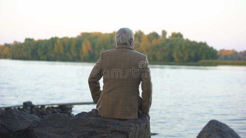 Séance masculine âgée sur la berge en pierre, pensant à la vie, appréciant la vue de nature banque de vidéos