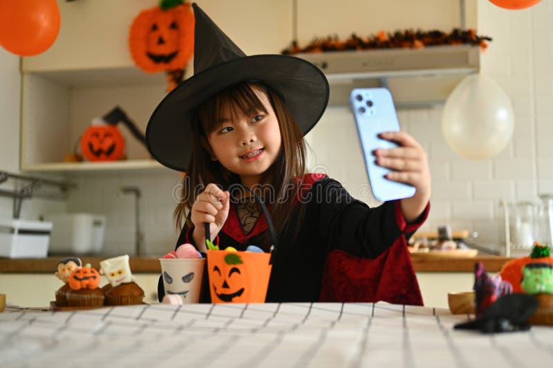 Sömmande liten flicka i häxkonst som visar halloween dessert och tar selfig med smartphone royaltyfri foto