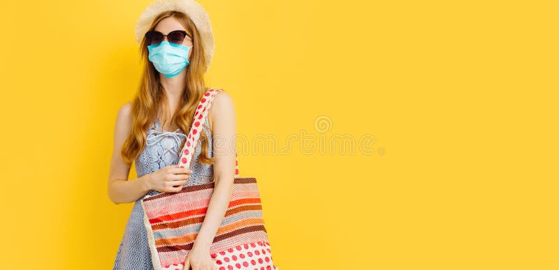 Sömmande ung kvinna i medicinmask i sommarklänning och hattställning medan man står och tittar på kameran över gul bakgrund royaltyfria bilder