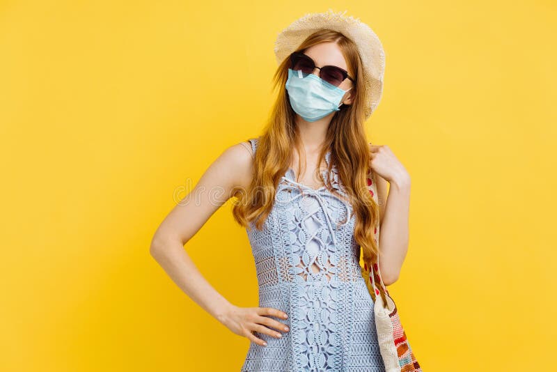 Sömmande ung kvinna i medicinmask i sommarklänning och hattställning medan man står och tittar på kameran över gul bakgrund royaltyfria foton