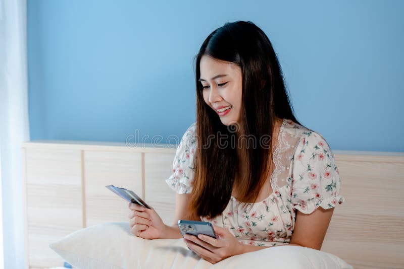 Cute Asian woman looking at 3 credit cards to choose to use for online shopping with the laptop in front of her bedroom.with a happy smiling face. Cute Asian woman looking at 3 credit cards to choose to use for online shopping with the laptop in front of her bedroom.with a happy smiling face.