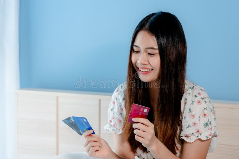 Cute Asian woman looking at 3 credit cards to choose to use for online shopping with the laptop in front of her bedroom.with a happy smiling face. Cute Asian woman looking at 3 credit cards to choose to use for online shopping with the laptop in front of her bedroom.with a happy smiling face