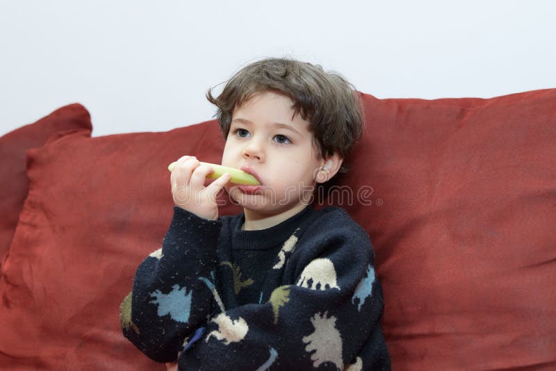 Söt babypojke som äter en gurka på soffan i sin dinasaur-pyjamas royaltyfri fotografi