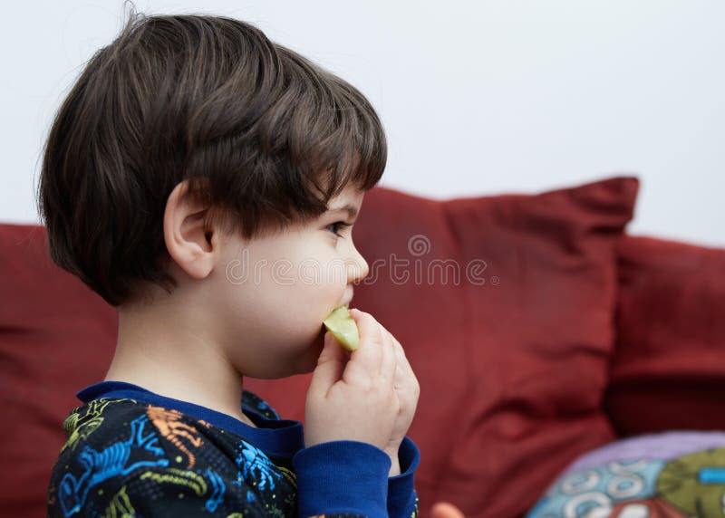 Söt babypojke som äter en gurka på soffan i sin dinosauripyjamas arkivfoto