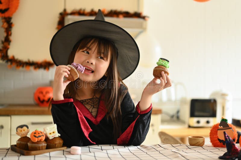 Söt liten flicka i häxkonst som står bakom köksbordet med halloweendesserter royaltyfri foto