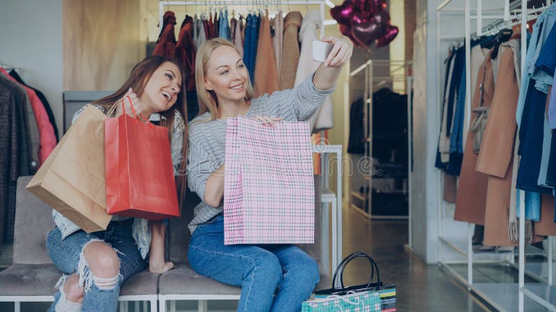 Söta flickor gör sig själviska med färgstarka papperspåsar i livmoderkläder, med smartphone och tittar på royaltyfria foton
