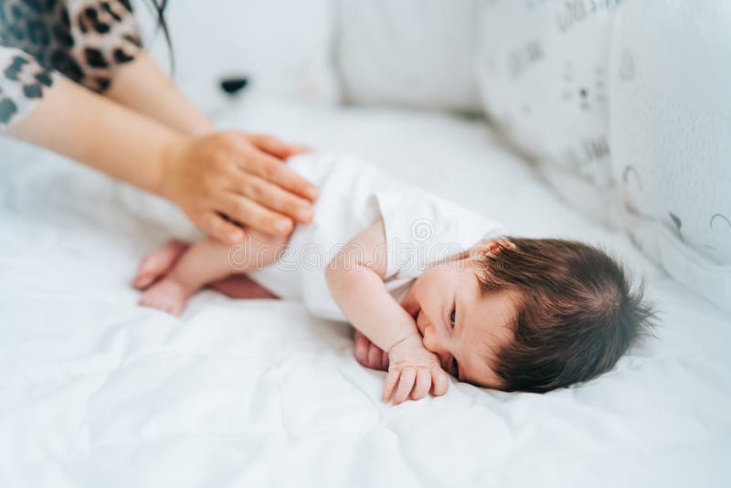 Cute little newborn baby with dark hair in white bodysuit lie in bed smiling and mother touching his legs. Healthcare, love, Mothers day, nursing, relationship concept. Selective focus. Copy space. Cute little newborn baby with dark hair in white bodysuit lie in bed smiling and mother touching his legs. Healthcare, love, Mothers day, nursing, relationship concept. Selective focus. Copy space.