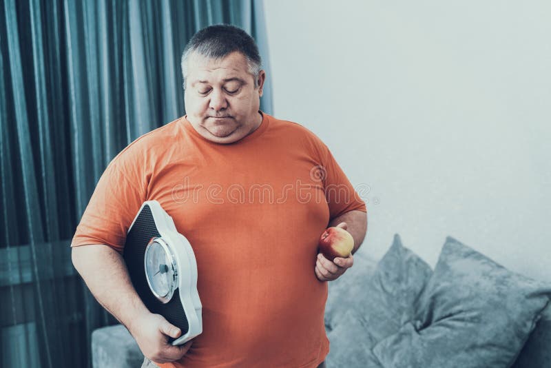 Fat Man i T-shirt with Weighter and Apple in Hands. Man with Bulimia. Unhealthy Lifestyle Concept. Sitting Man. Sad Man. Treatment of Overweight. Man with Overweight. Healthy and Vegetarian Food. Fat Man i T-shirt with Weighter and Apple in Hands. Man with Bulimia. Unhealthy Lifestyle Concept. Sitting Man. Sad Man. Treatment of Overweight. Man with Overweight. Healthy and Vegetarian Food.