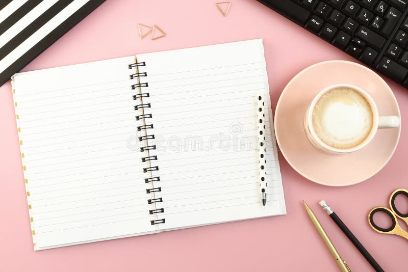 Tabela cor-de-rosa da mesa de escritório com caderno, a xícara de café, a pena, o lápis, as tesouras e o computador abertos