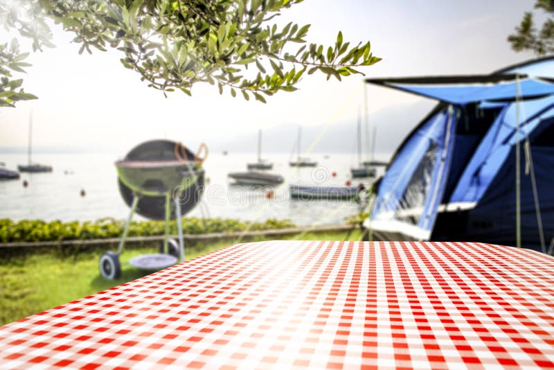 Red and white checkered tablecloth on the table in garden with blurred lake background and space for your decoration. Grill with smoke on grass. Red and white checkered tablecloth on the table in garden with blurred lake background and space for your decoration. Grill with smoke on grass.